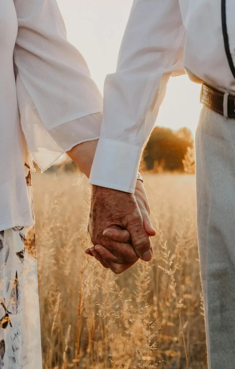 An elderly couple holding hands as they walk together through a golden field at sunset